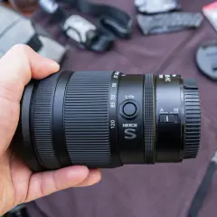 Dłoń trzyma obiektyw Nikkor S 24-120mm, idealny do pełnej klatki Nikon. W tle akcesoria fotograficzne.
