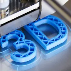 Drukarka 3D tworzy niebieskie litery "3D" z plastiku.